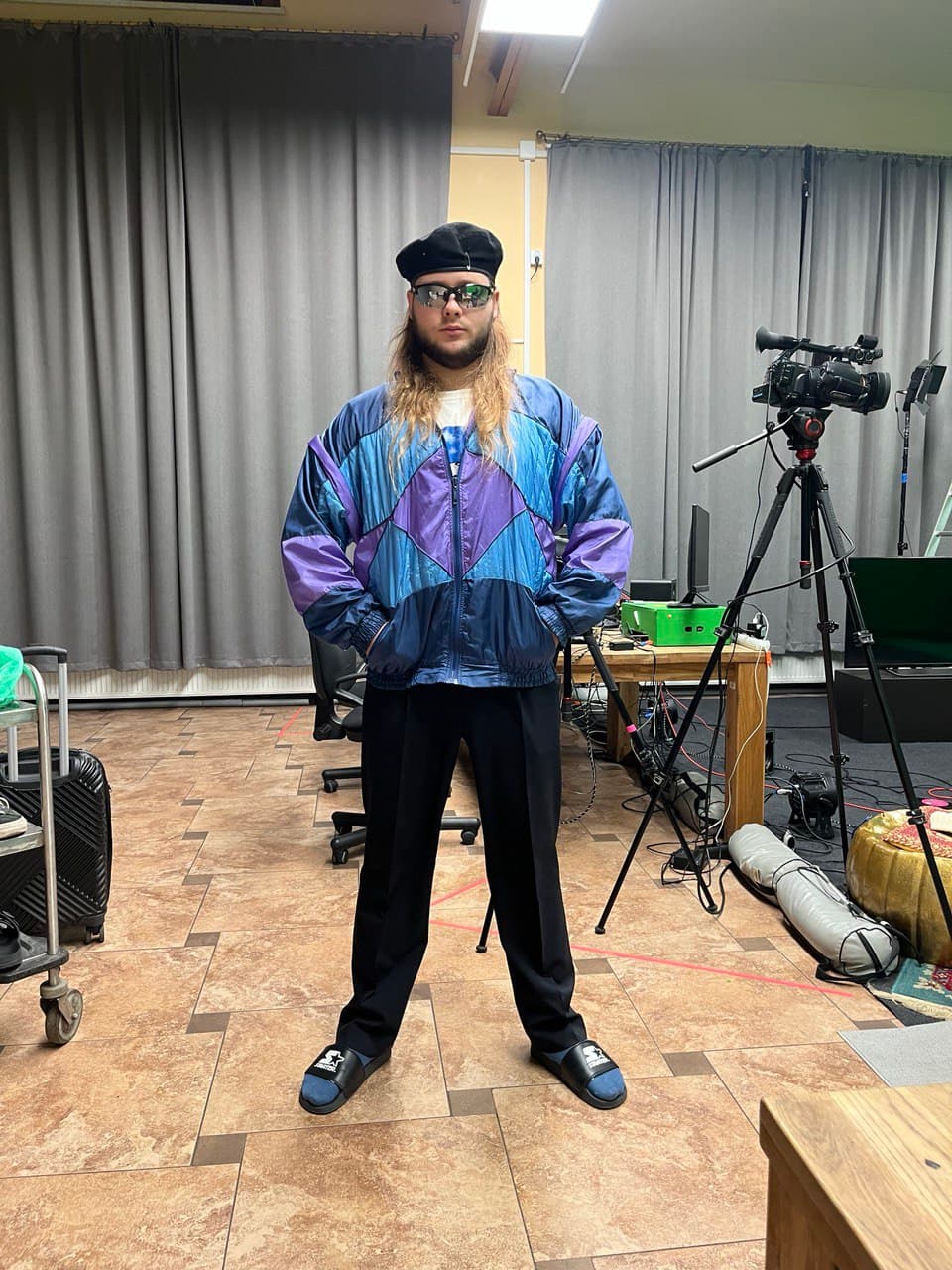 Hlib Dovzhuk in a production studio, wearing a vintage windbreaker jacket, sunglasses, and a beret, standing among broadcast camera equipment